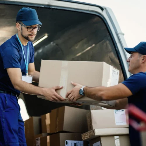 Dos repartidores de mensajería cargando un paquete de cartón en la parte trasera de una camioneta de entregas.
